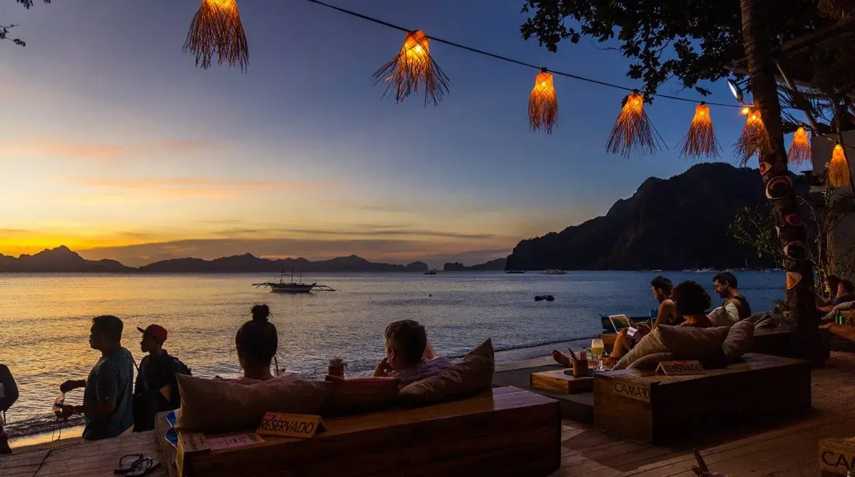 Baie au crépuscule avec des lanternes allumées, des tables basses et des canapés en bois au beach club