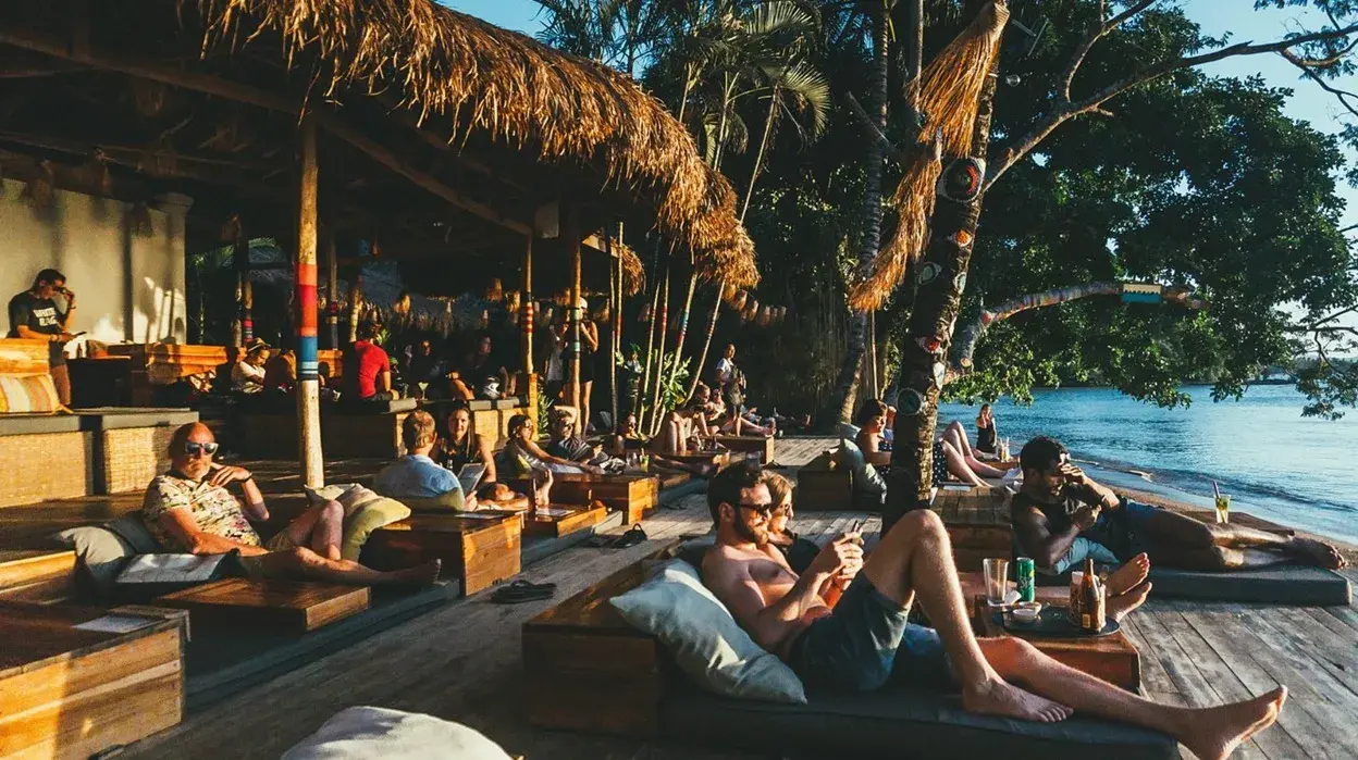 Terrasse du beach club avec un toit de chaume, des lits-canapés en bois et des clients qui discutent au bord de la mer
