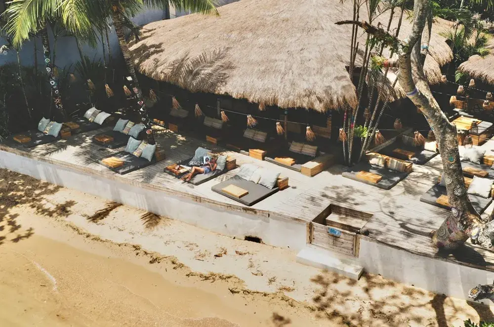 Vue en hauteur de la terrasse en bois du beach club avec des transats, un toit de chaume et le sable de la plage