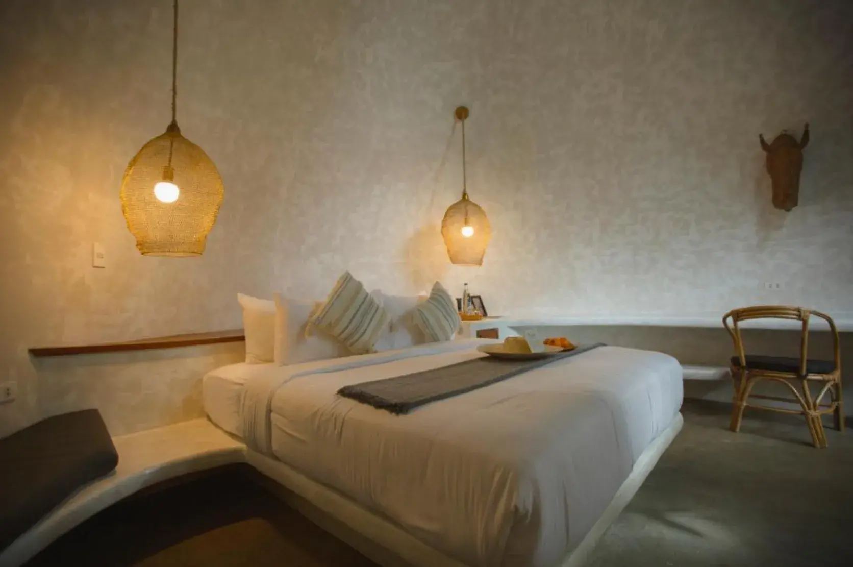 Chambre avec un lit en linge blanc, deux lampes en filet à la lumière chaude et une chaise en rotin