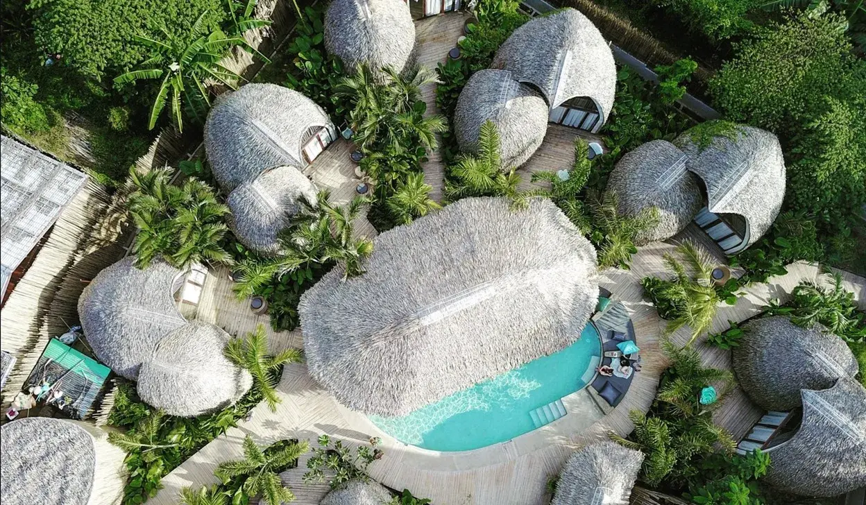 Vue aérienne de la piscine turquoise et des transats, entourés de dômes au toit de chaume et de palmiers